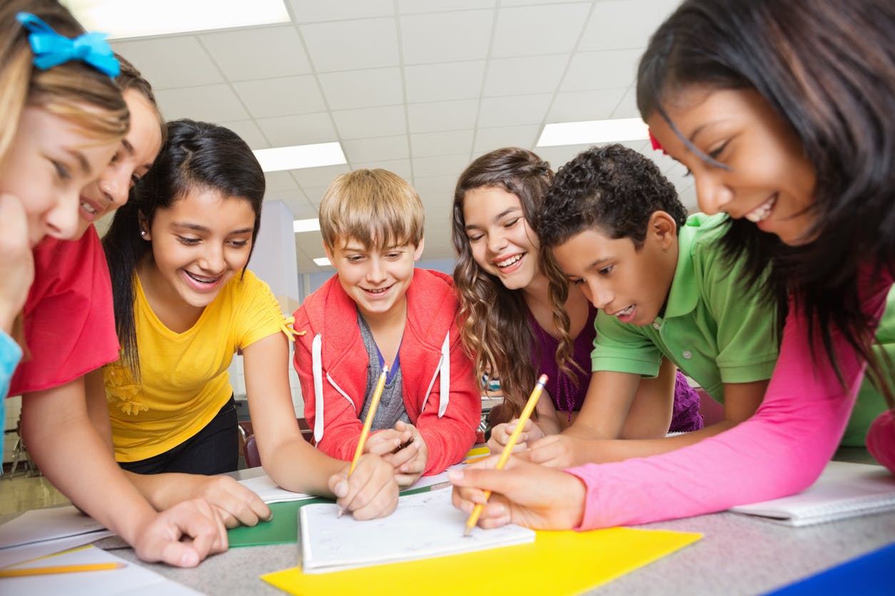 Seven middle school students gather around a table smiling while doing a writing exercise.