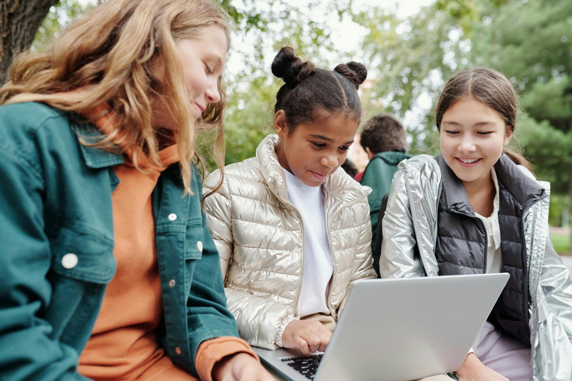 Three young girls sit outside in a park and work on a laptop as they learn online together.