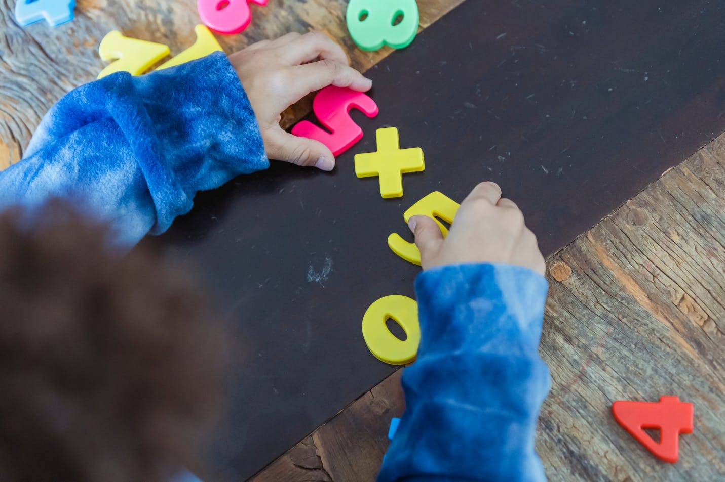 Child using movable numbers and math symbols on a table to show a 5x5 formula and help someone else improve their math skills