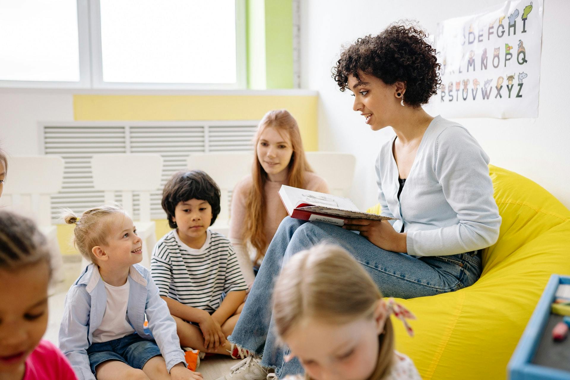 A young teacher reads a story to a group of students sitting on the ground.