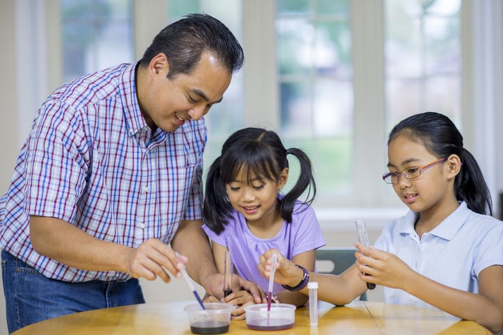 A father helps his two young daughters with an at-home science experiment.