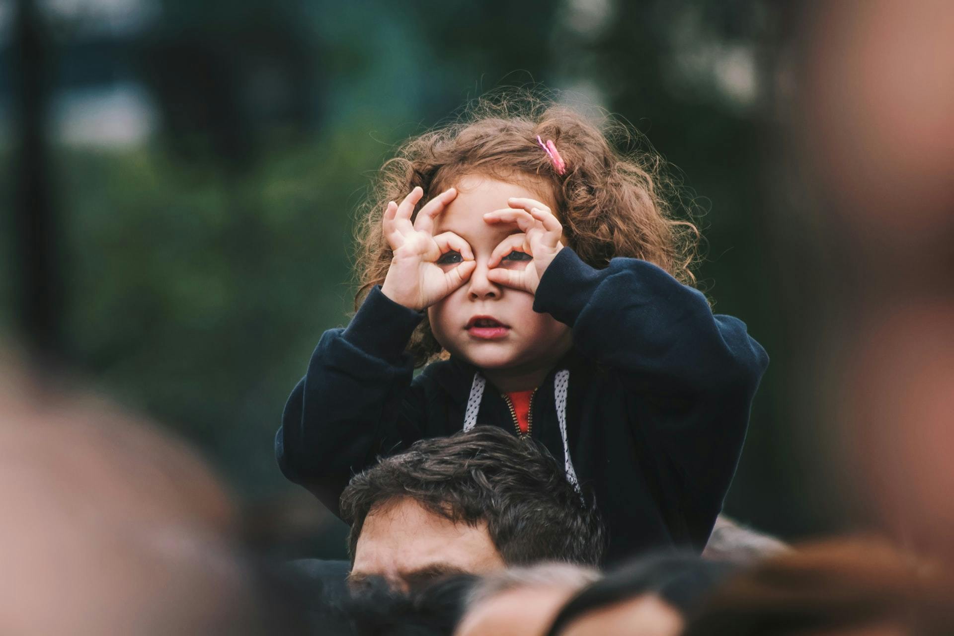 Young girl sits on her father's shoulders and peers through her fingers atop a crowd. 