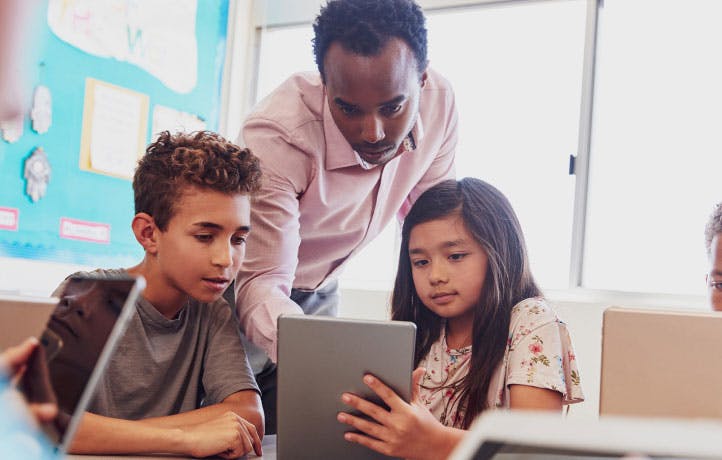 A teacher guides two students playing math and English games on tablets.
