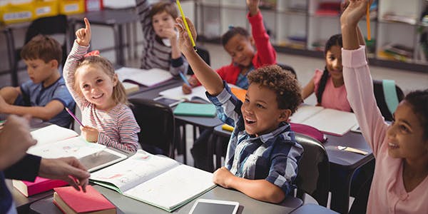 Young students sit in rows, raising their hands to answer the teacher's question.