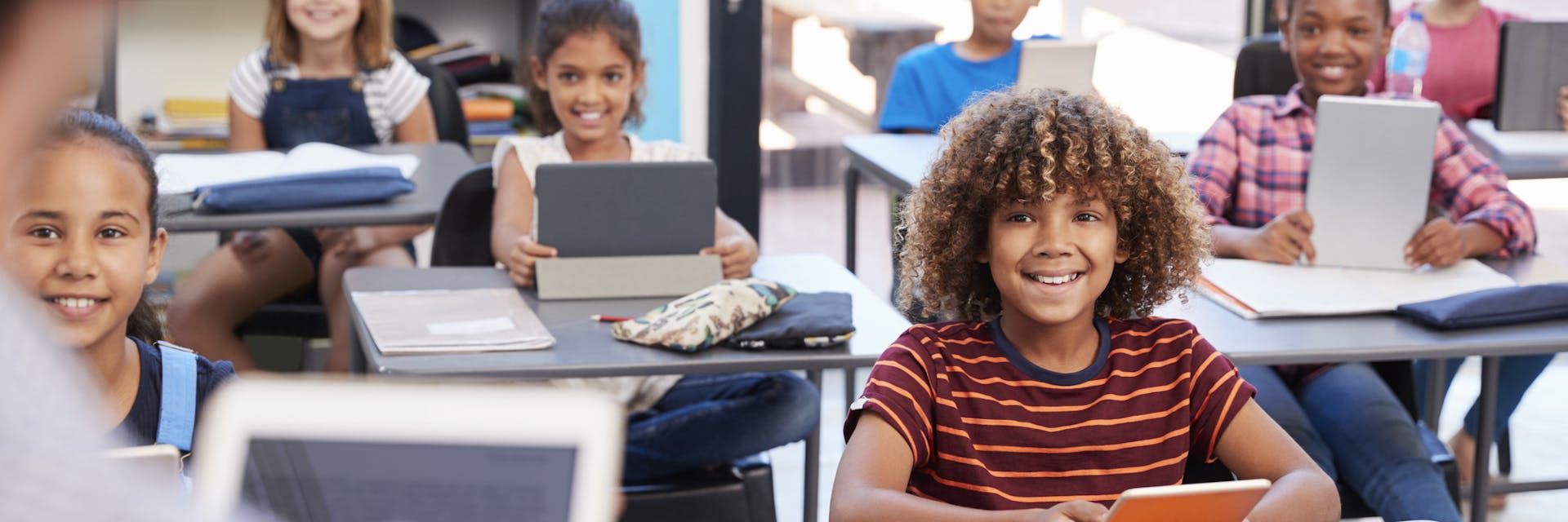 A classroom of smiling children sitting at desks and holding tablets faces the teacher at the front of the room. 