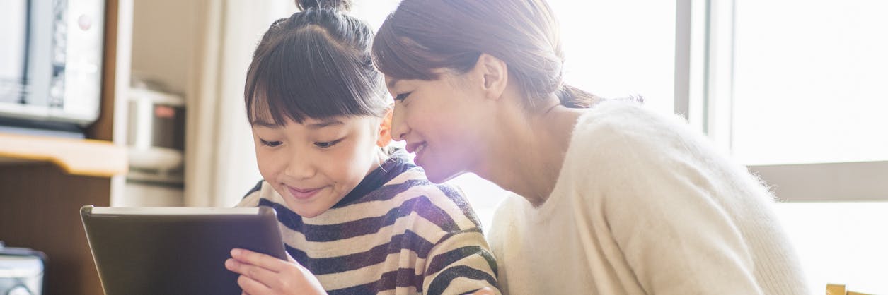 A mother looking at what her daughter is playing on a tablet at kitchen table.