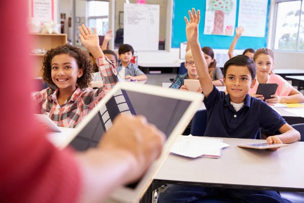 Teacher stands at the front of a classroom holding a tablet while her class of students raise their hands