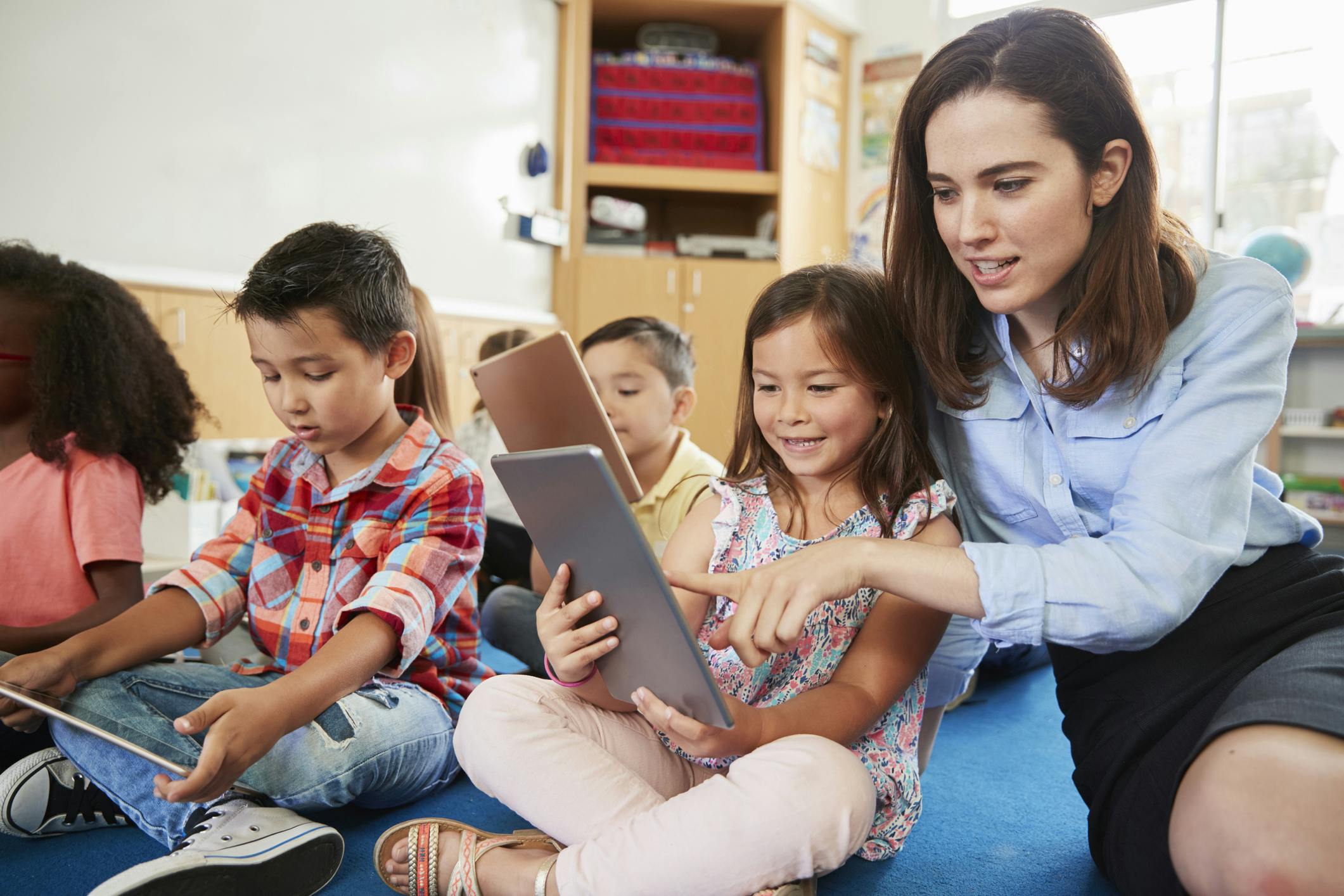 Teacher works with group of children sitting on the floor and playing Prodigy on iPads.