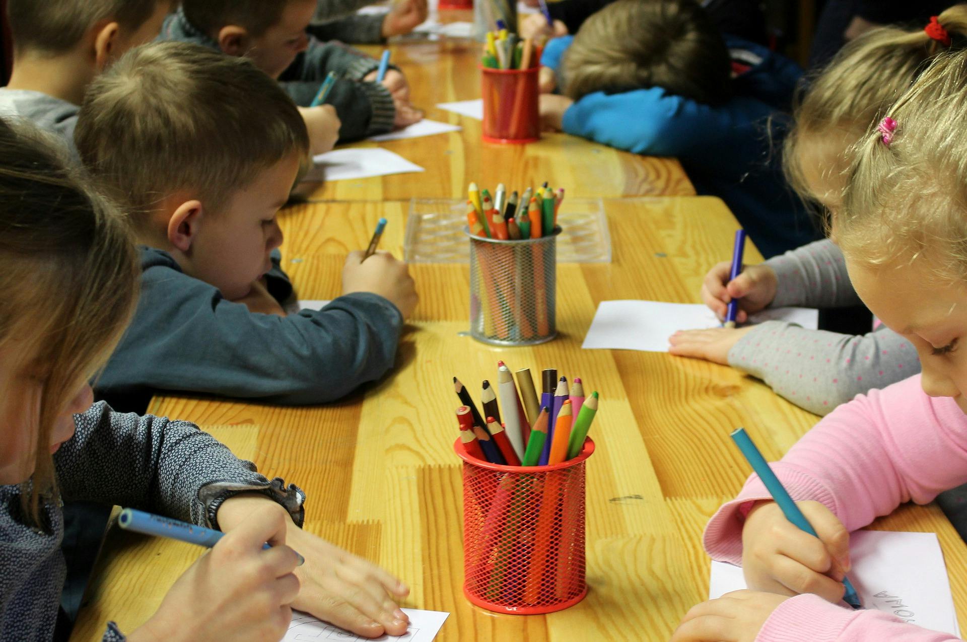 A group of kindergarten students sits at a long table, writing on paper with pencil crayons.
