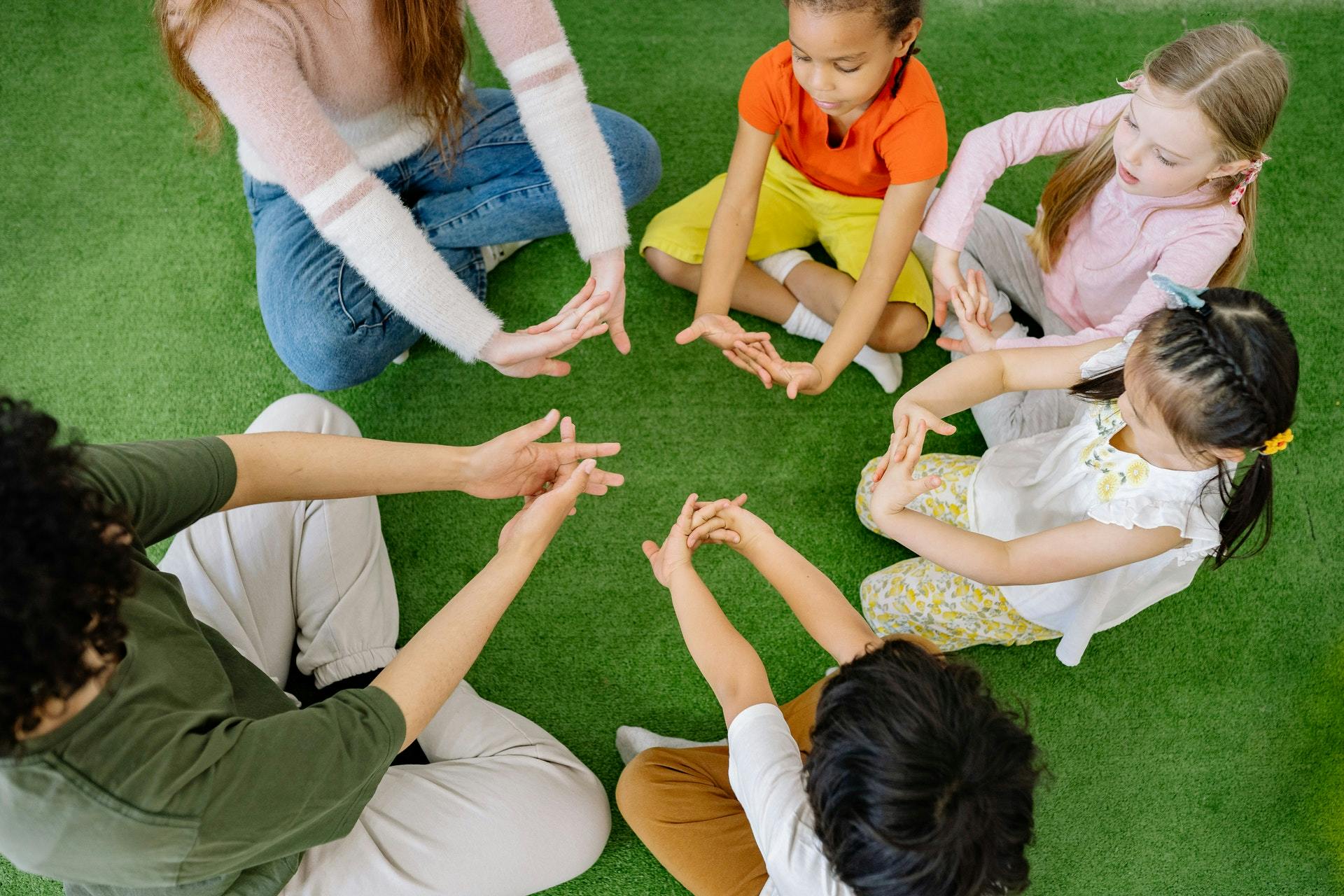 Four young students and two teachers sit in a circle and stretch their hands.