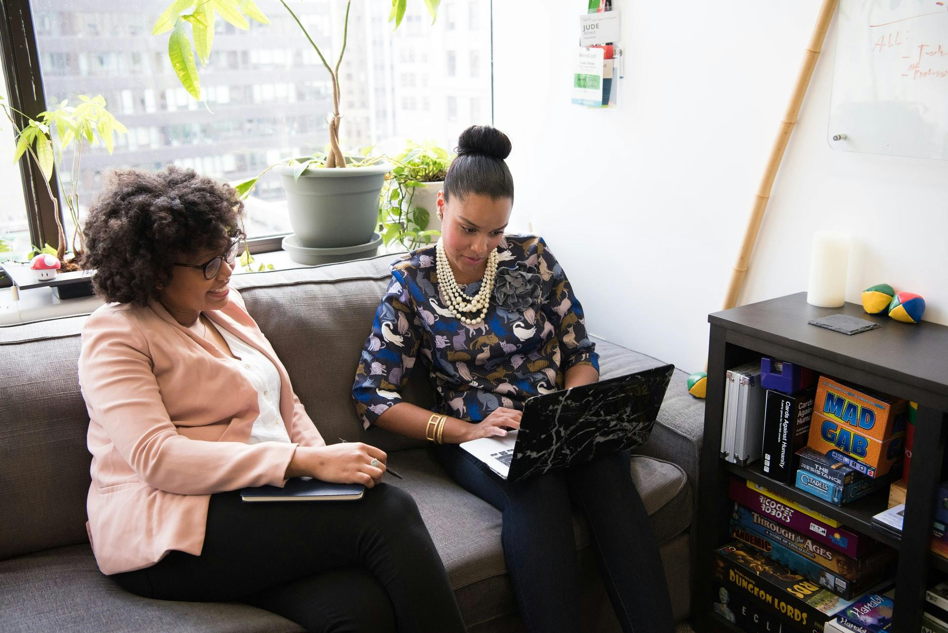 Two teachers sitting on a couch in front of a window and discussing something on a laptop screen.