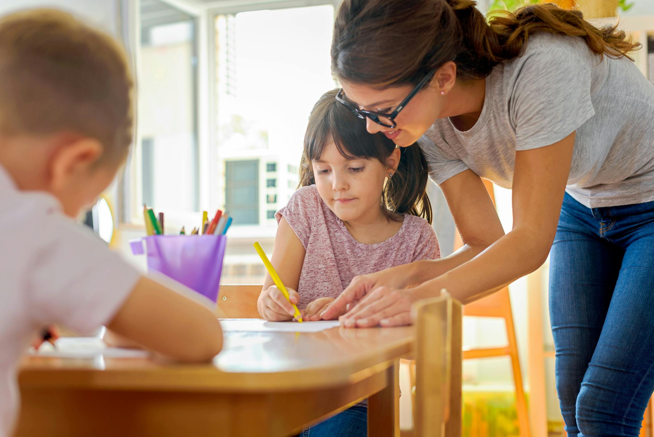 A teacher works one on one with a kindergarten student working at a wooden table.