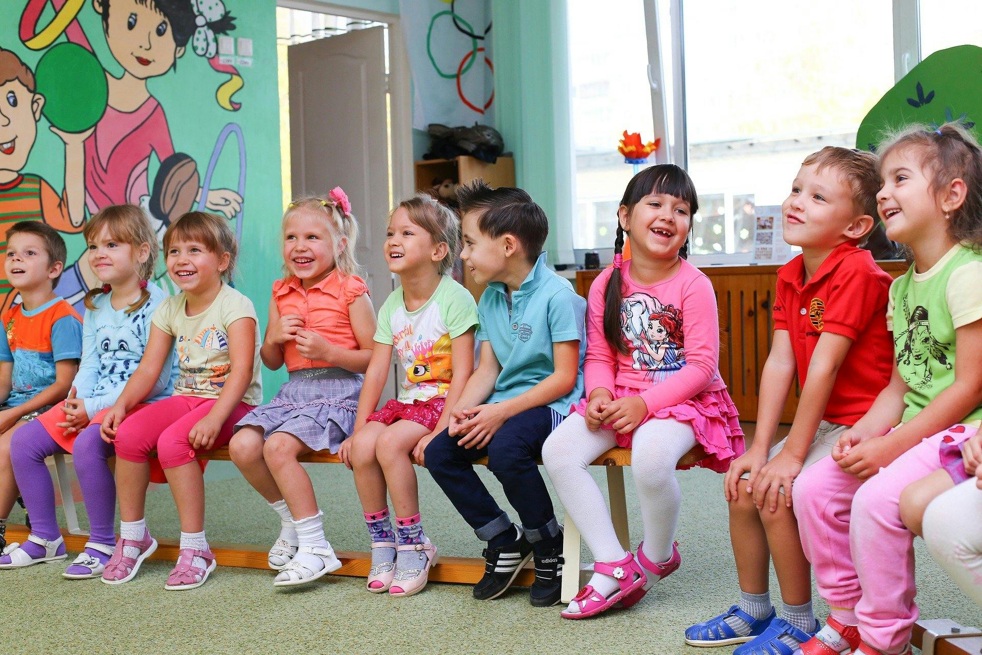 Nine kindergarten students sit in a row on wooden benches in a classroom.