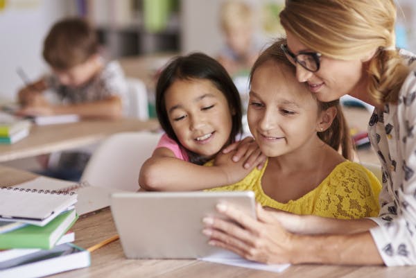 A teacher in her classroom shows two students educational content on a tablet.