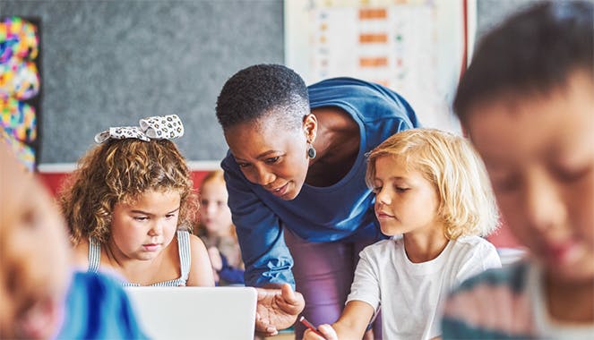 A classroom teacher pointing at a computer, showing her two students how to use Prodigy to practice math skills from their curriculum