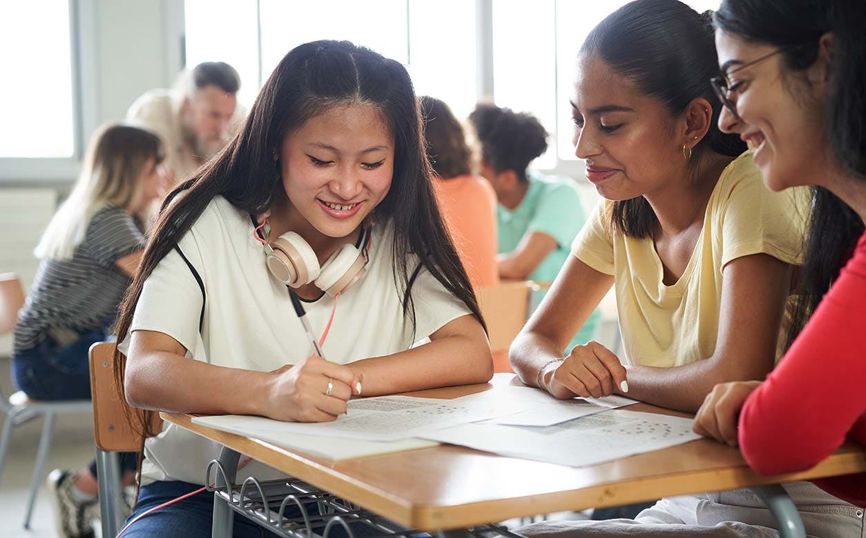 Three students sit at a table, engaged in conversation while looking at a notebook in front of them.