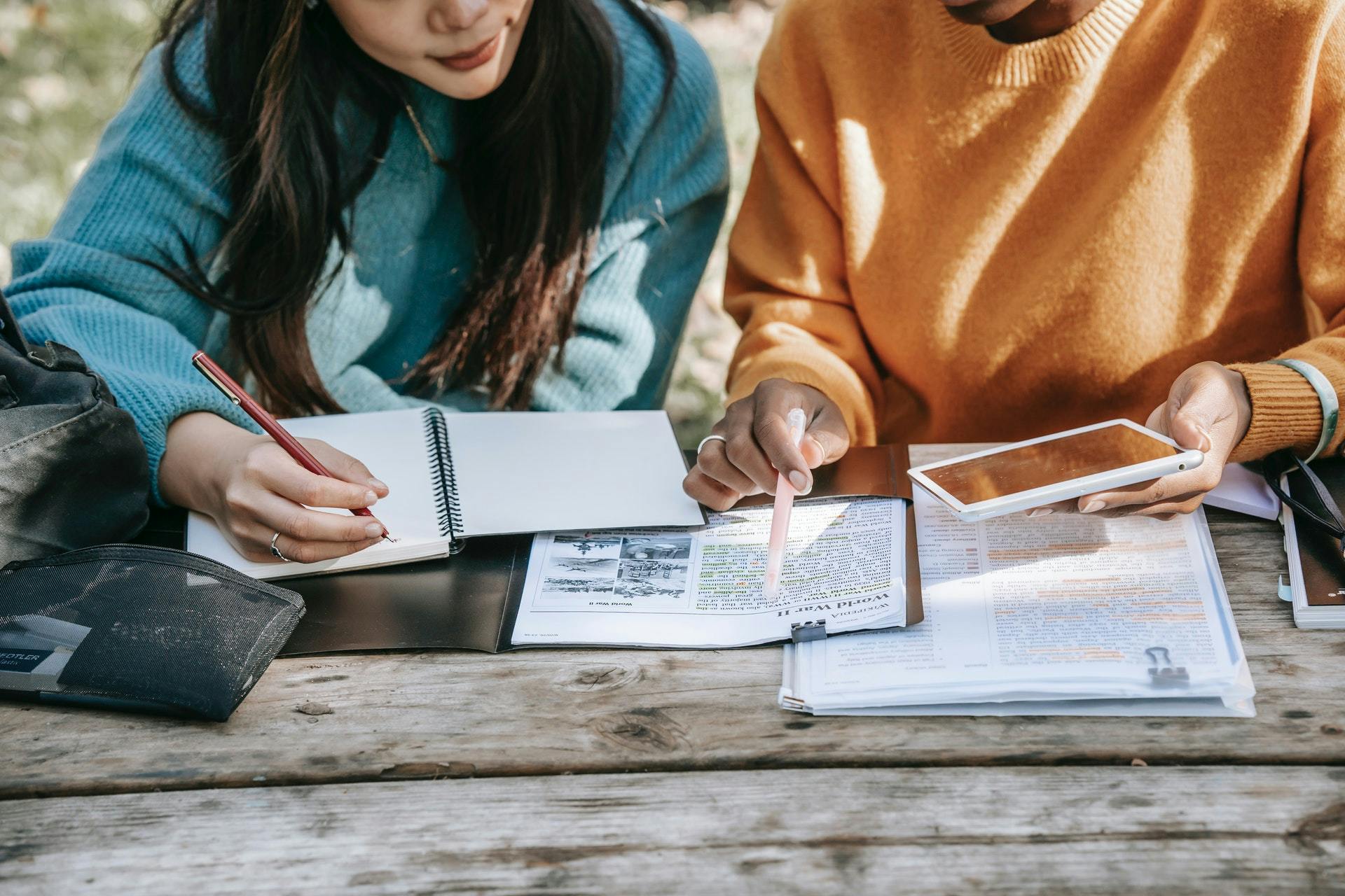 Two students sit at a table outside and study while using a learning app.