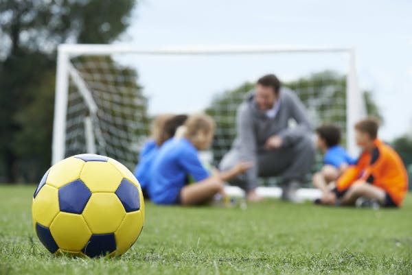 Close-up of a yellow and blue soccer ball while a PE teacher and his four students talk near a soccer net.