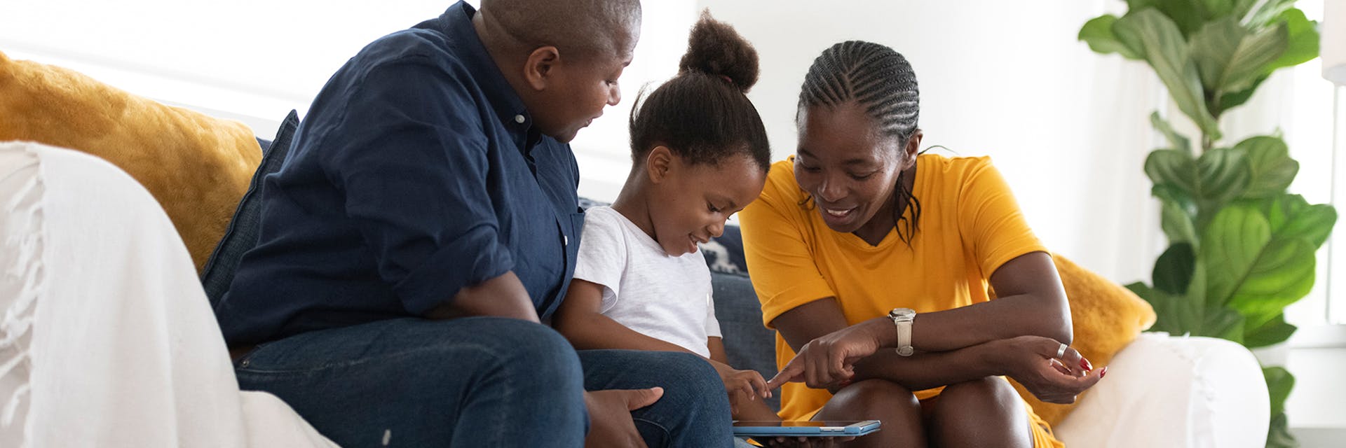 Two parents and their daughter sit on the couch and look at a device together.