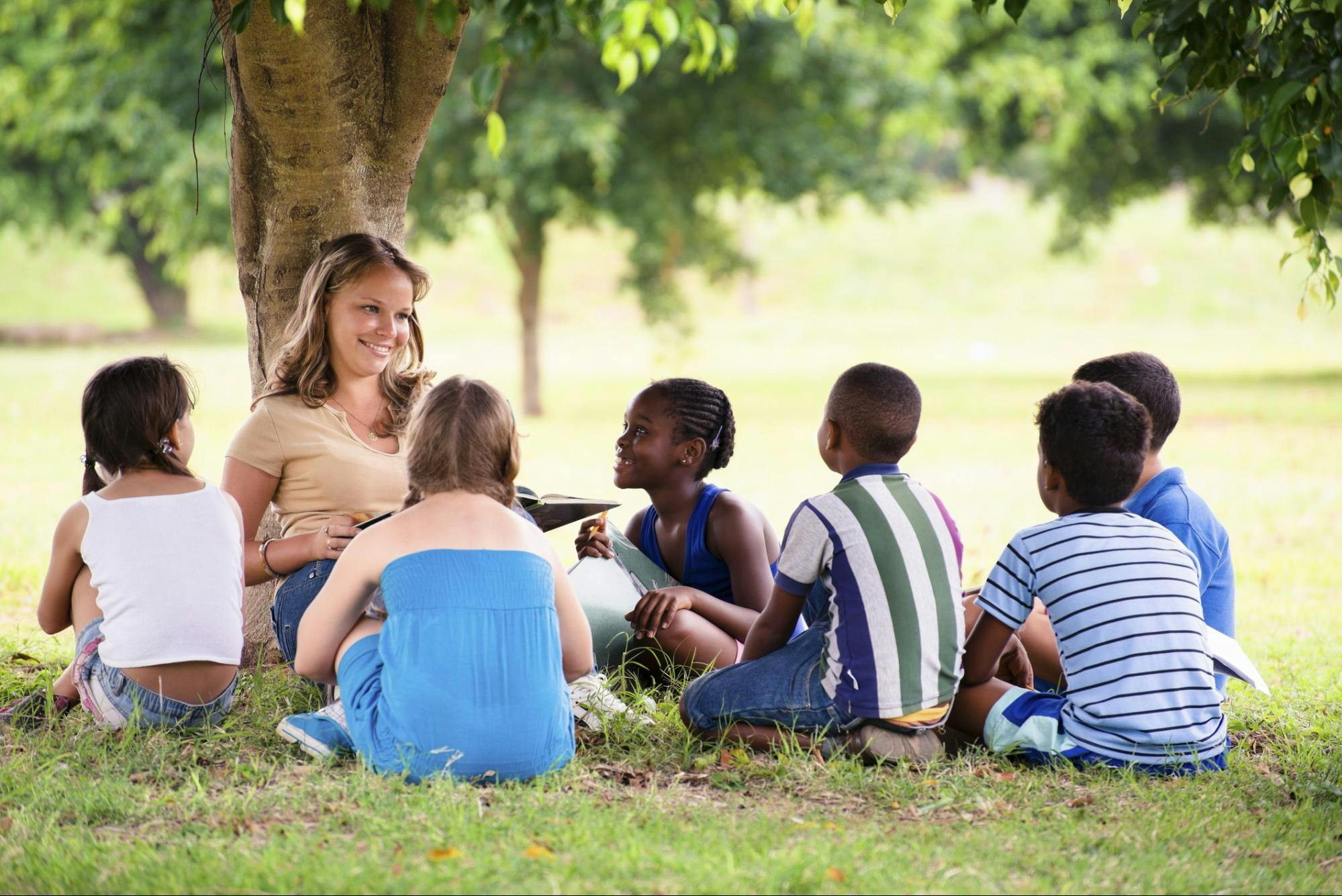 A teacher and a group of students sit outside underneath a tree.