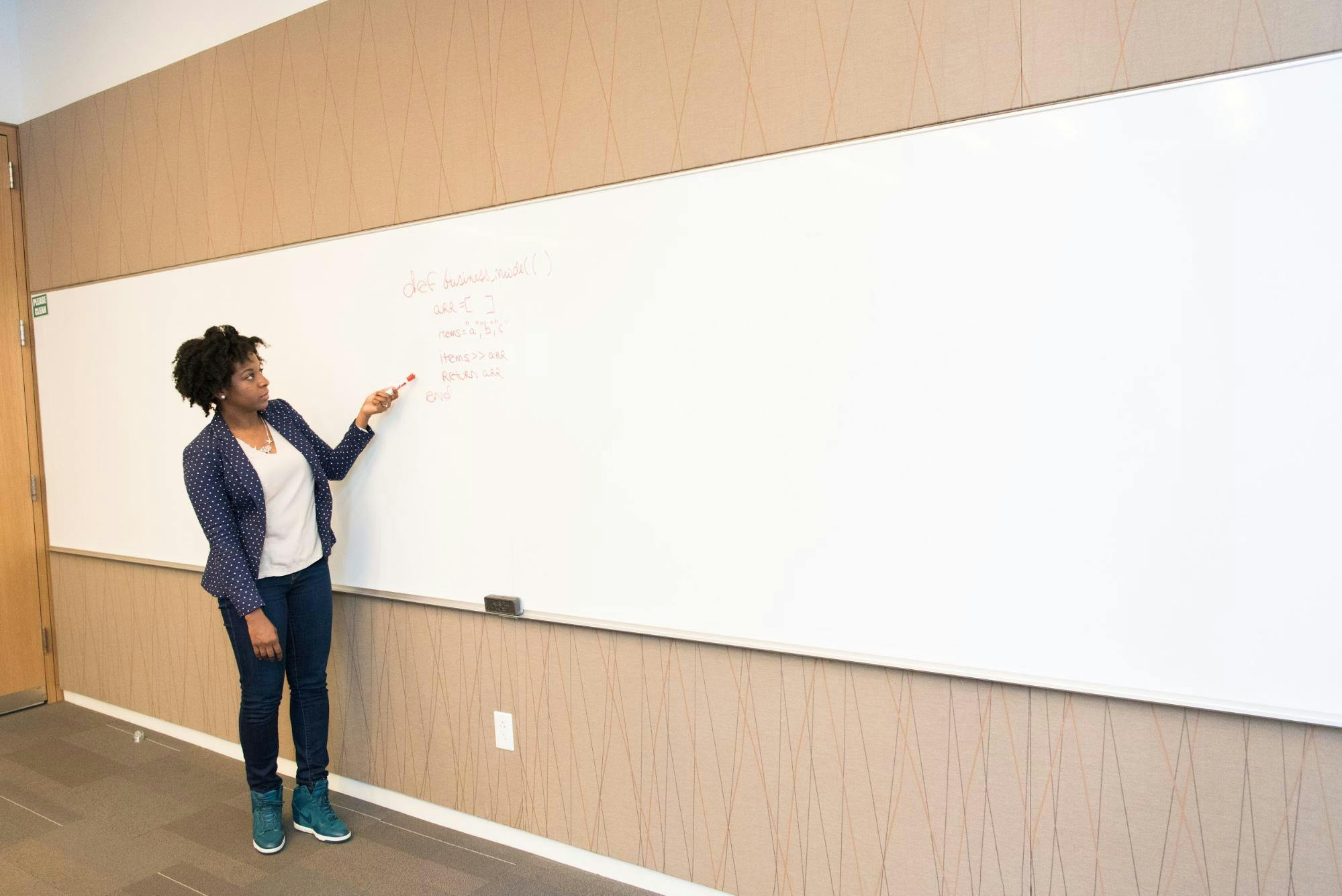 Teacher stands at whiteboard and points with marker at red writing.