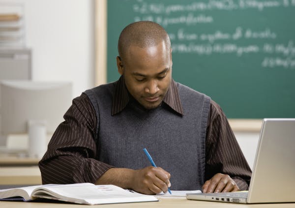 Teacher grading papers in a classroom with a pen while having a textbook and laptop nearby.