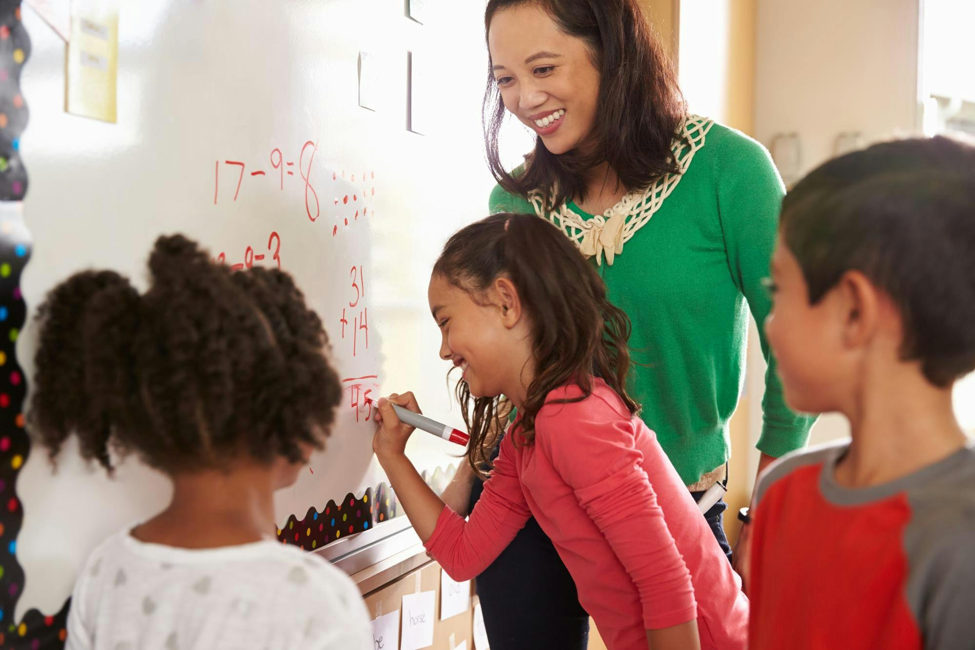 A teacher smiles at three students working on a math problem on a whiteboard.