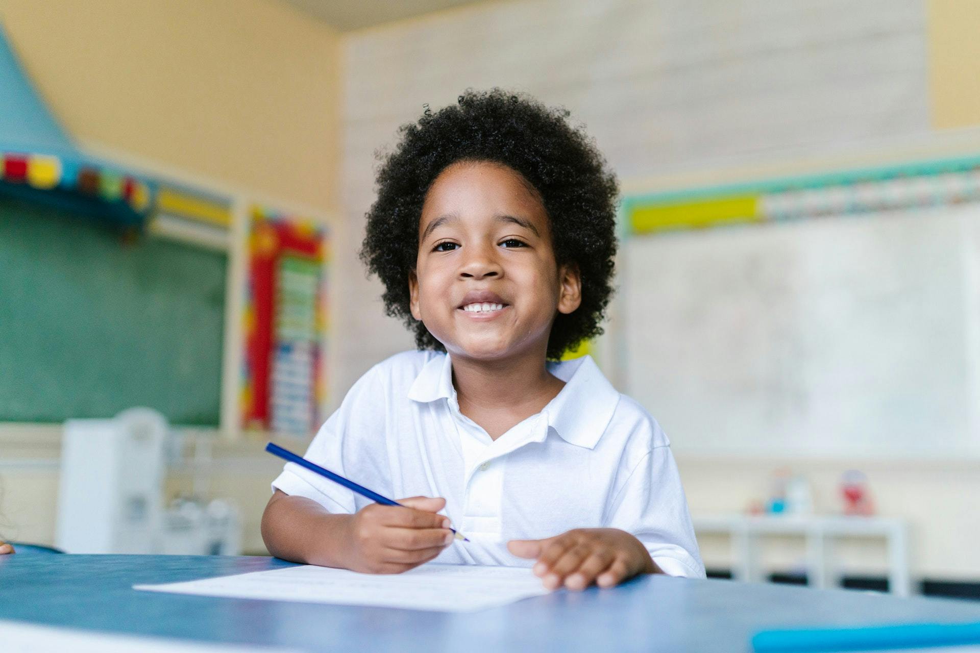 Young boy smiles while sitting at a table holding a pencil.