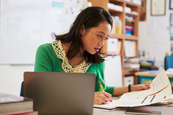 Teacher reviewing student performance on paper while having her laptop beside her.