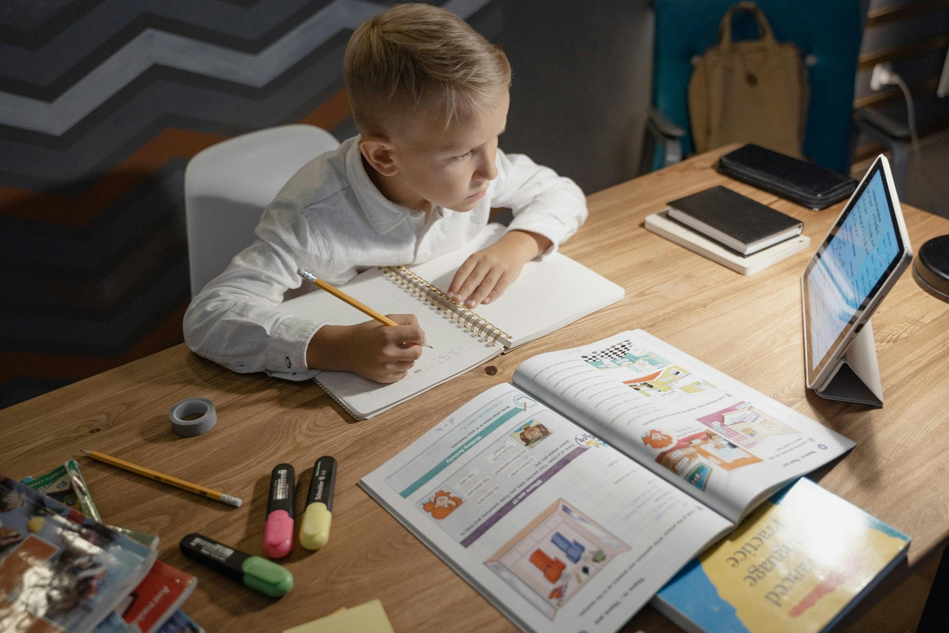 Young boy works at a desk with a tablet, notebook and workbook as he practices ELL skills. 