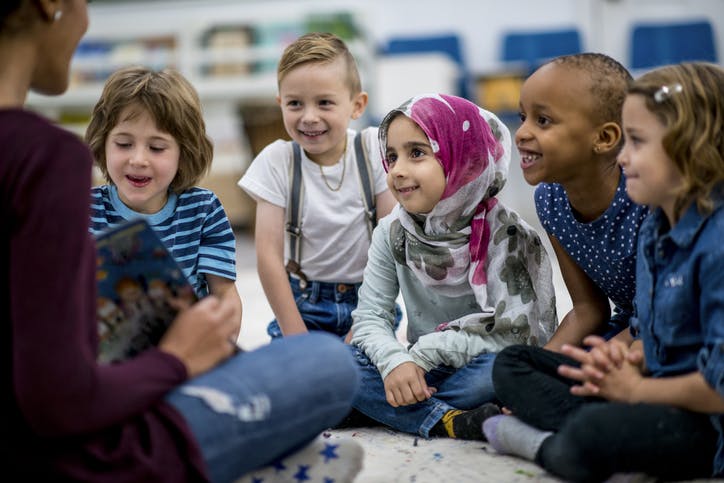 Five students sit cross legged on the ground in front of a teacher holding a book.