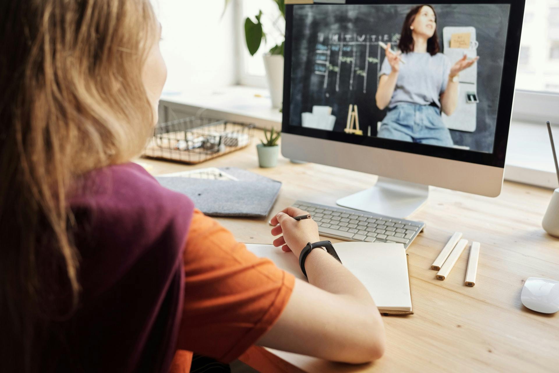 Student sits at at desk at home, writing in a notebook and watching a female teacher lecture in front of a blackboard on the computer in front of her.