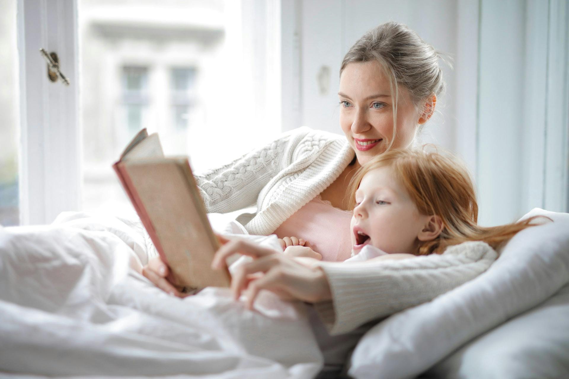 A mom and daughter lie in bed and read. 