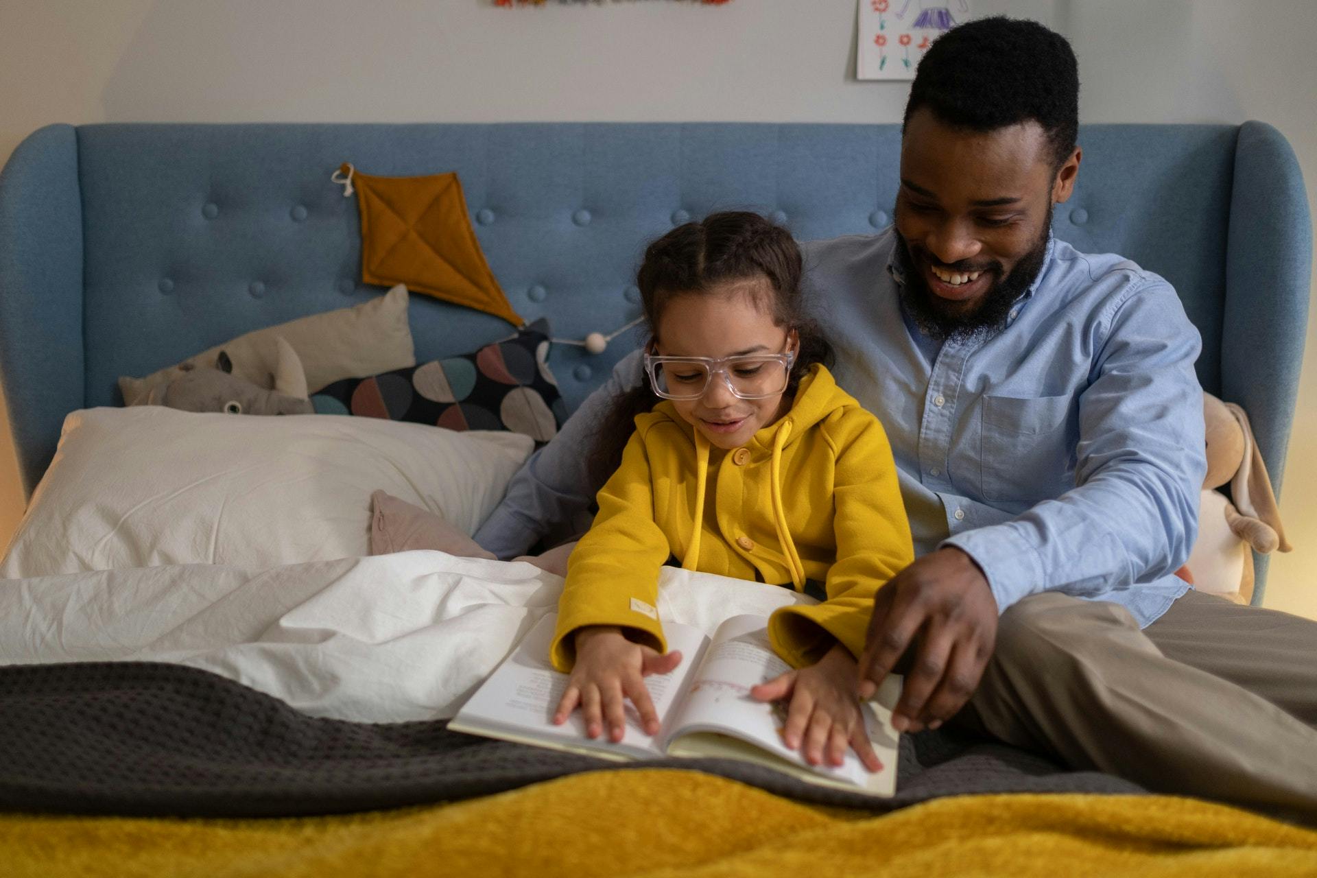 Father and daughter sit on a bed reading a book for first grade reading activities. 