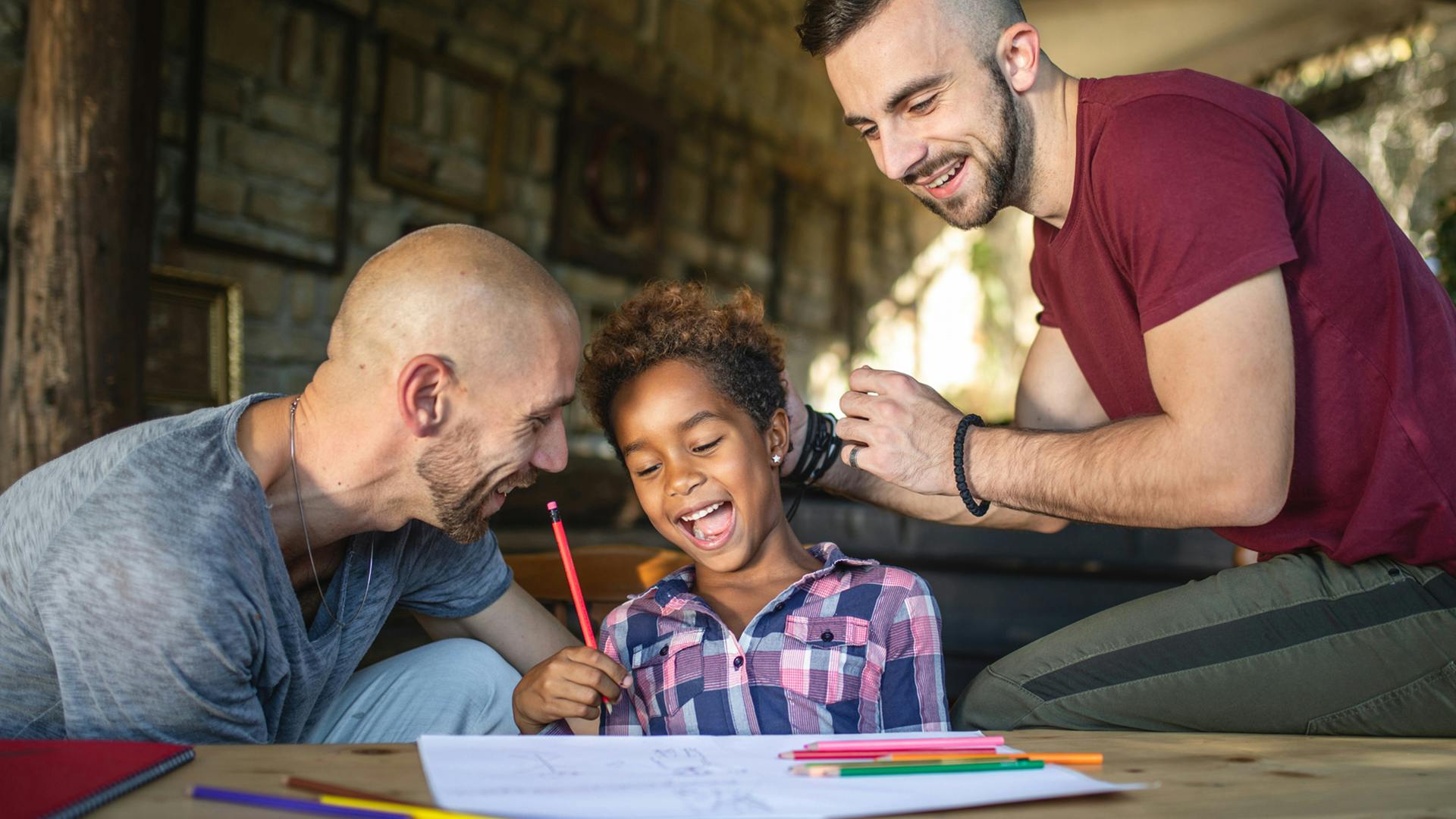 Two fathers and a young girl who is laughing work on completing a homework assignment at the table. 
