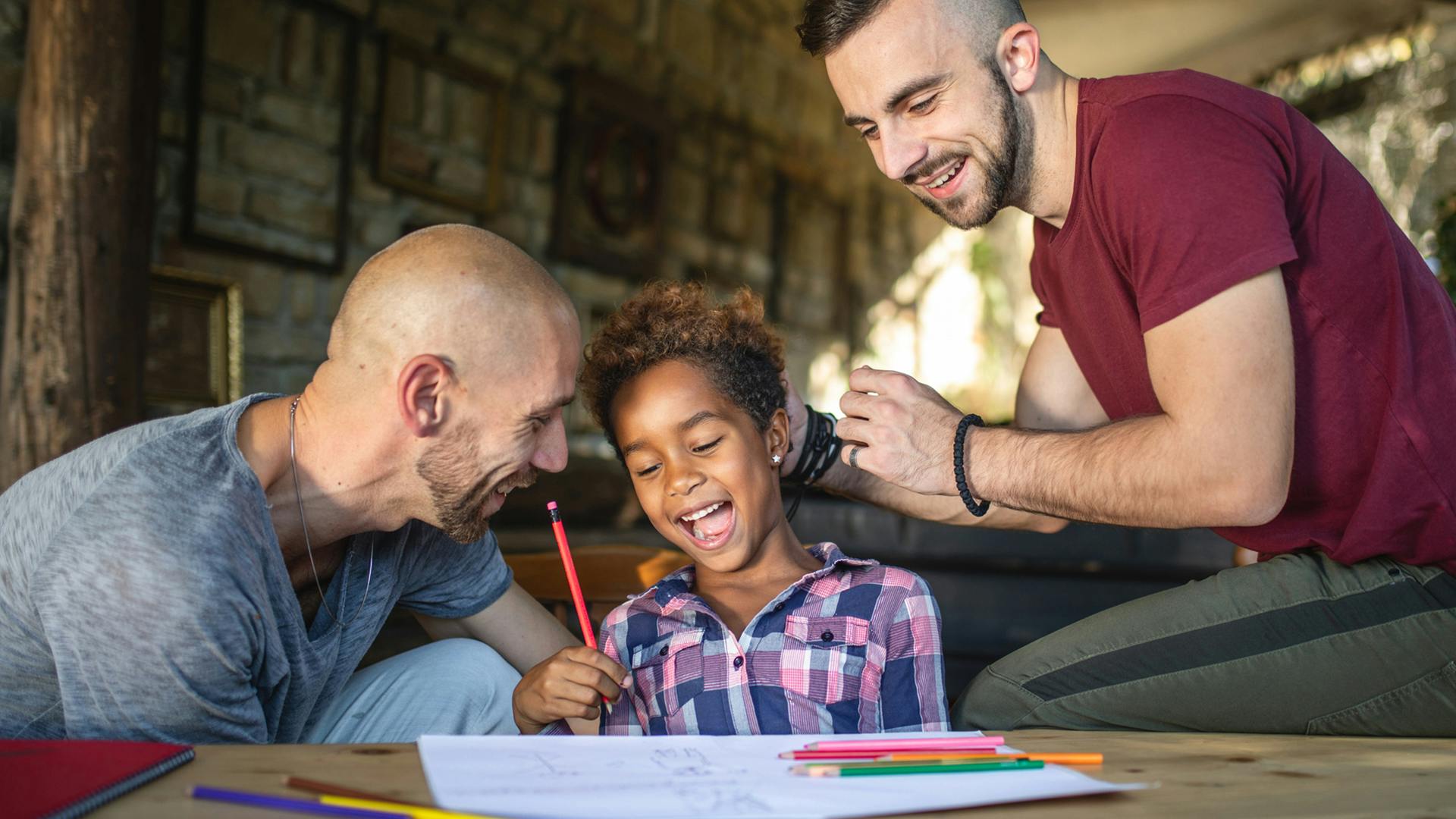 Two fathers and a young girl who is laughing work on completing a homework assignment at the table.