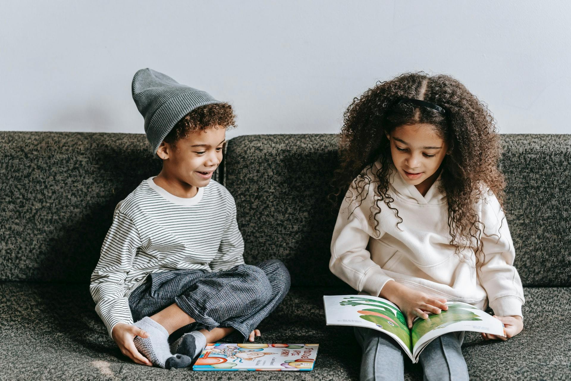 Two elementary aged children sit on a grey couch and read picture books for 1st graders.