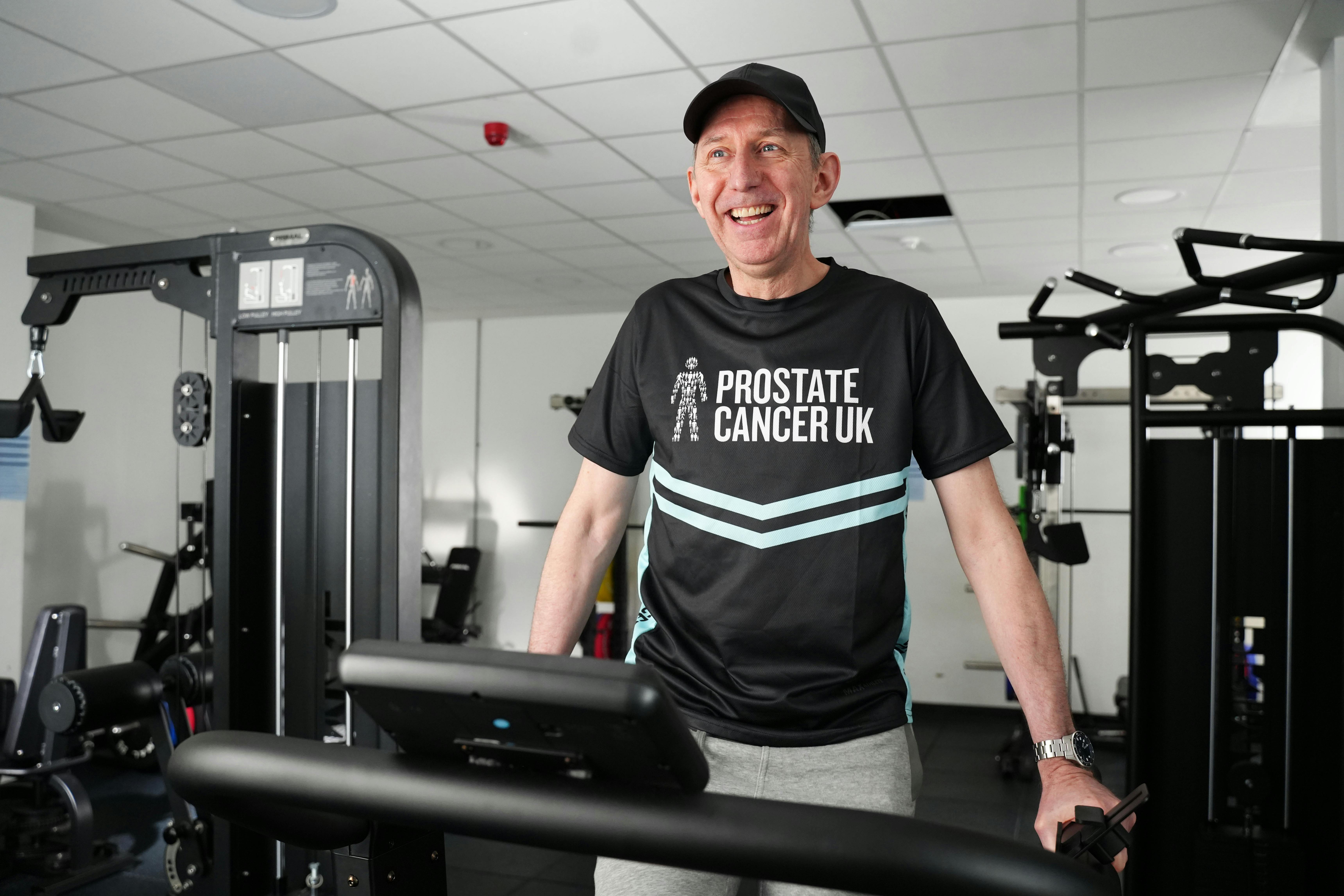A man smiling at the camera, standing on a treadmill wearing a cap and branded Prostate Cancer UK running t-shirt