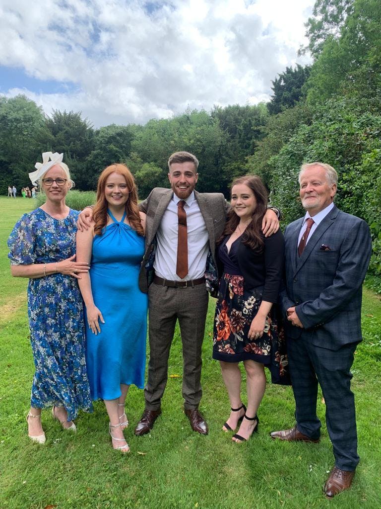 A family of 2 men and 3 women standing together surrounded by trees. 