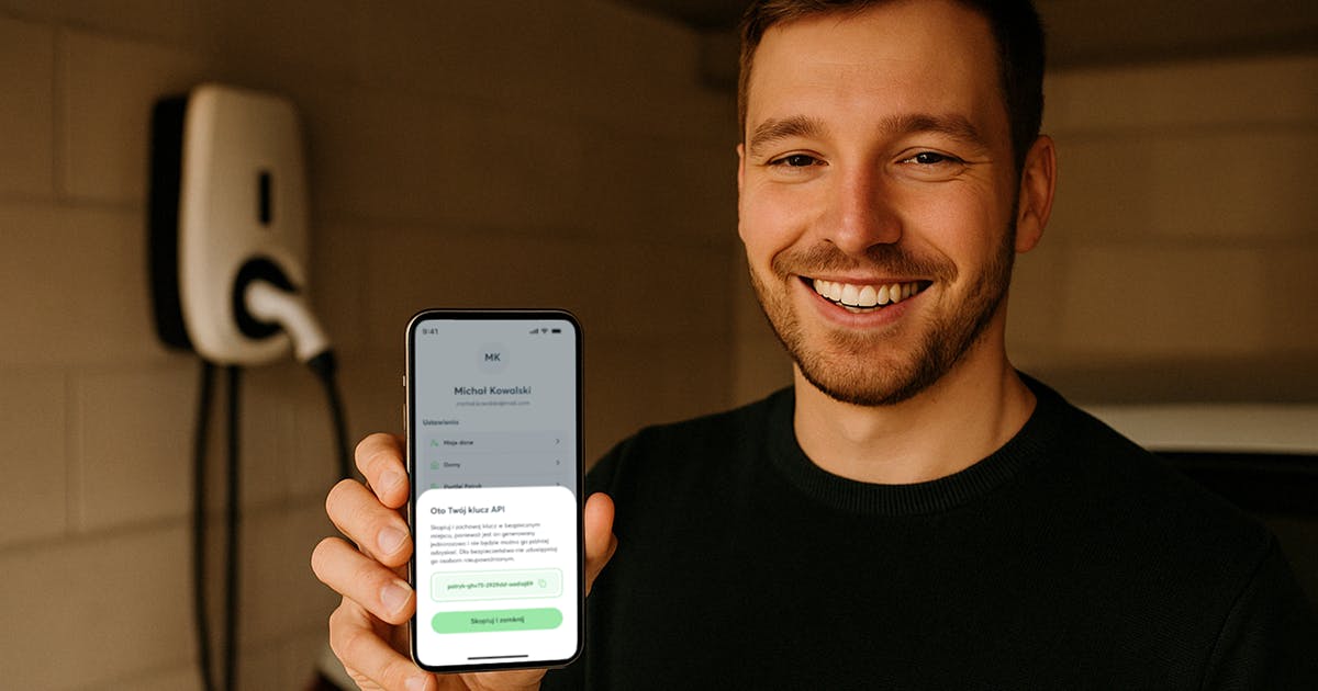 Mężczyzna trzymający w dłoni telefon z uruchomioną aplikacją Pstryk na tle garażu z ładowarką samochodu elektrycznego