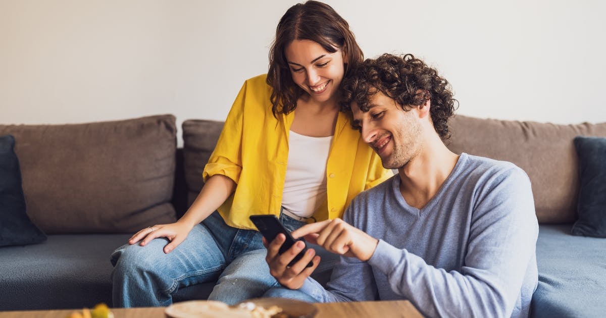 Para siedząca na kanapie, przeglądająca smartfona – domowa scena związana z opłacaniem rachunków online lub przeglądaniem faktury za prąd.