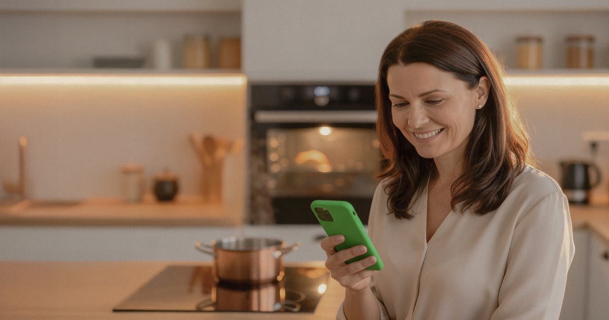 Uśmiechnięta kobieta z telefonem w dłoni, w tle działający piekarnik.