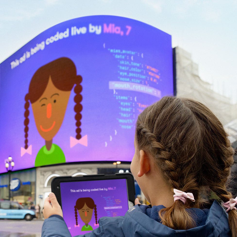 Young person holding an iPad displaying an avatar and computer code, the iPad display mirrored onto the large display at Piccadilly Circus.