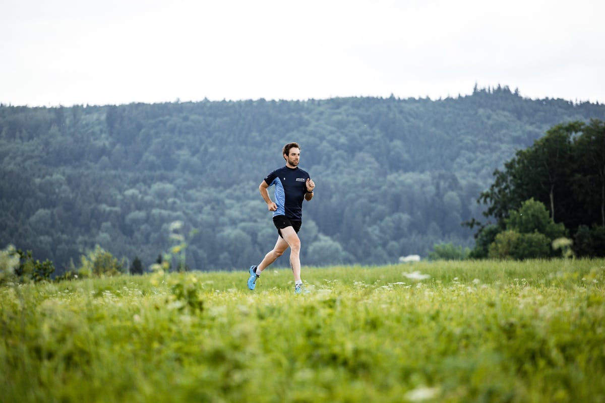 Ein Läufer im blauen Sportoutfit läuft über eine Wiese, dahinter eine Bergkulisse.