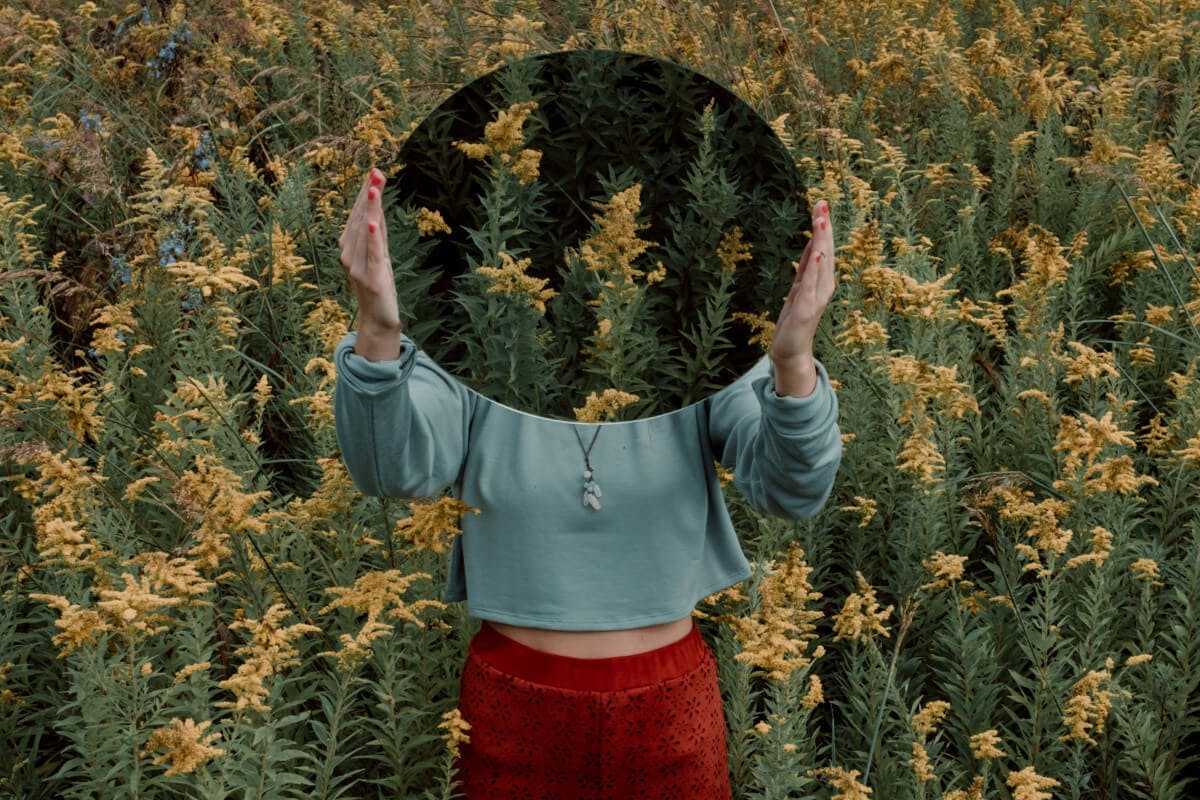 Frau steht in einem Blumenfeld und hält sich einen Spiegel vors Gesicht.