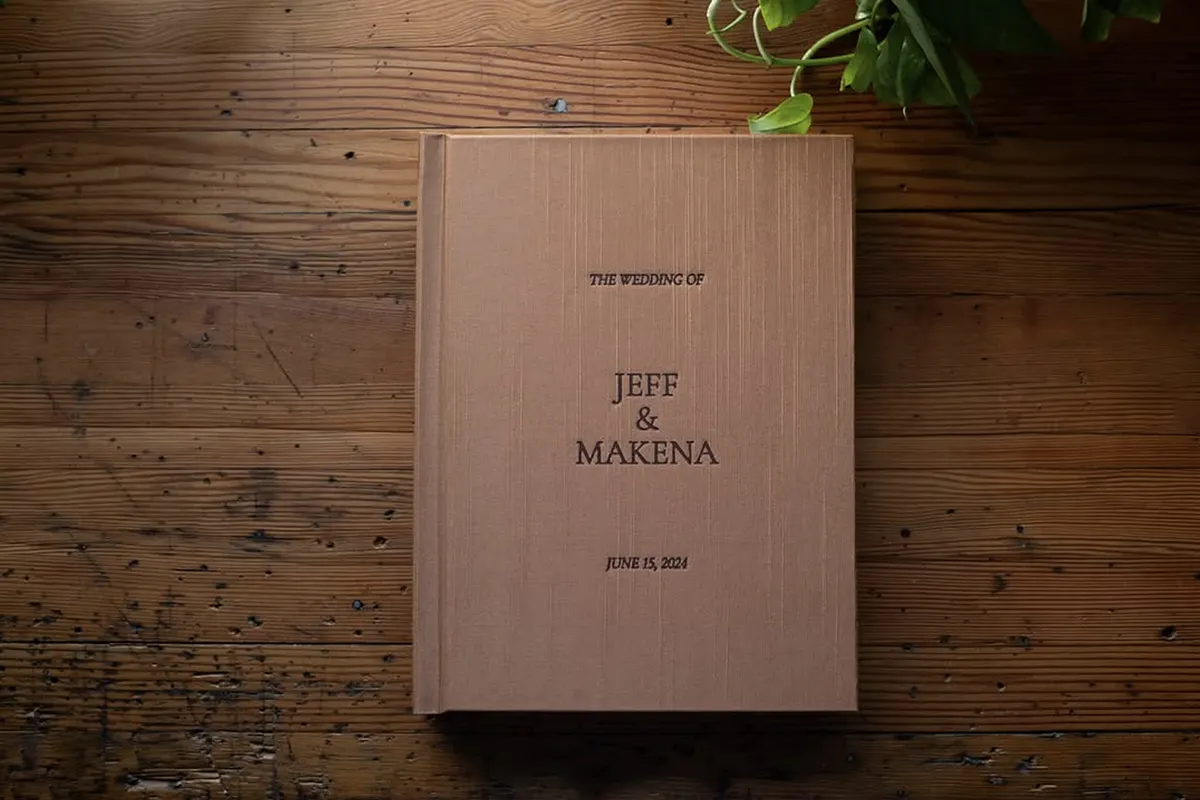 Vision Art mauve linen wedding album debossed with The Wedding of Jeff and Makena on a rustic wood table with greenery