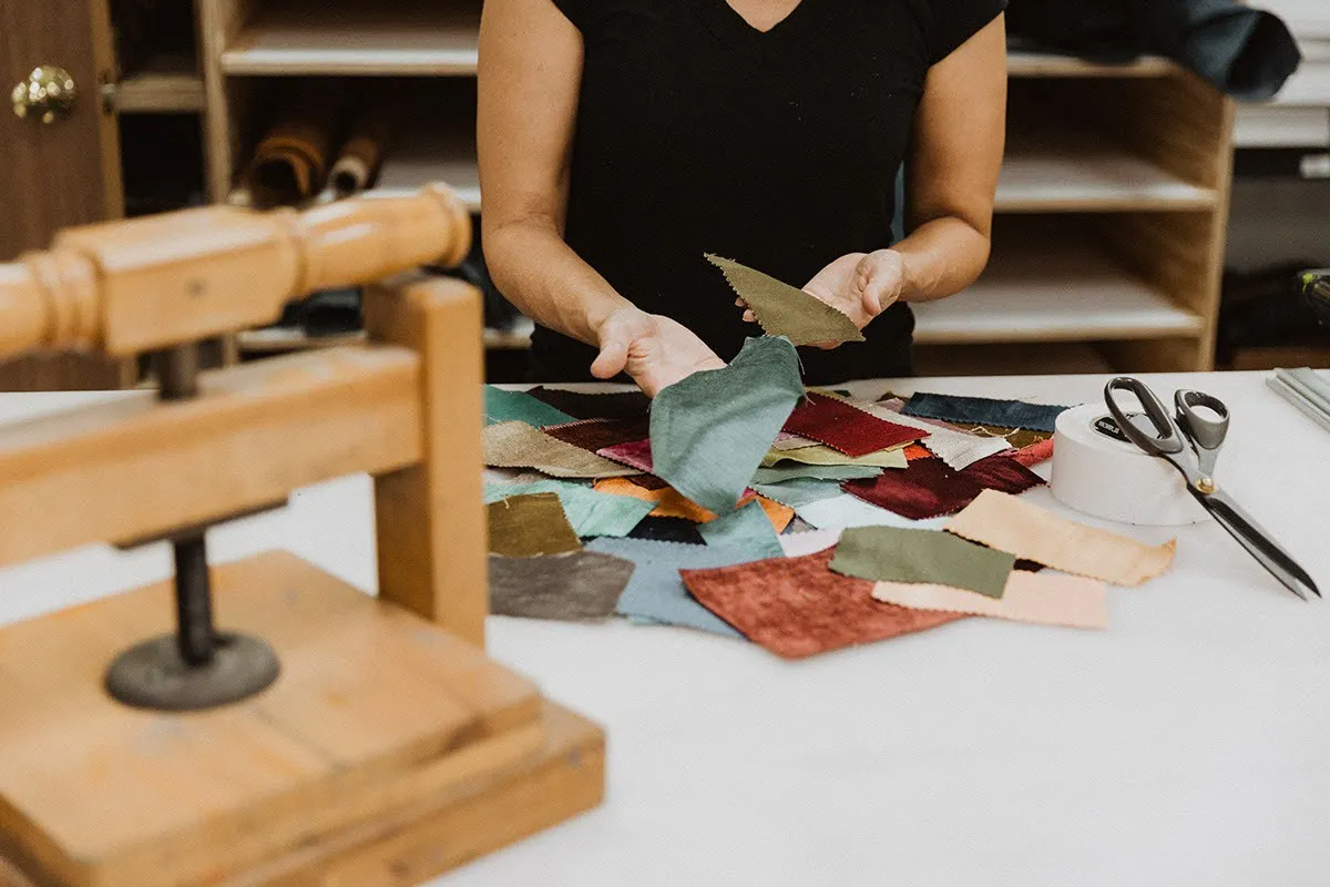 Vision Art artisan selecting colored linen fabric swatches at a bookbinding workbench with a wooden book press, scissors, and tape
