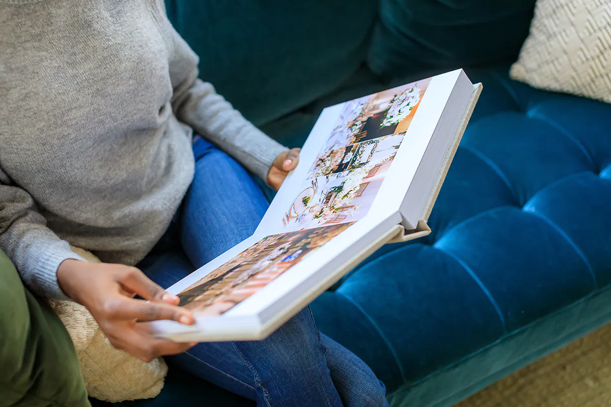Woman on a teal velvet sofa turning pages of an open Kiss Books wedding album.