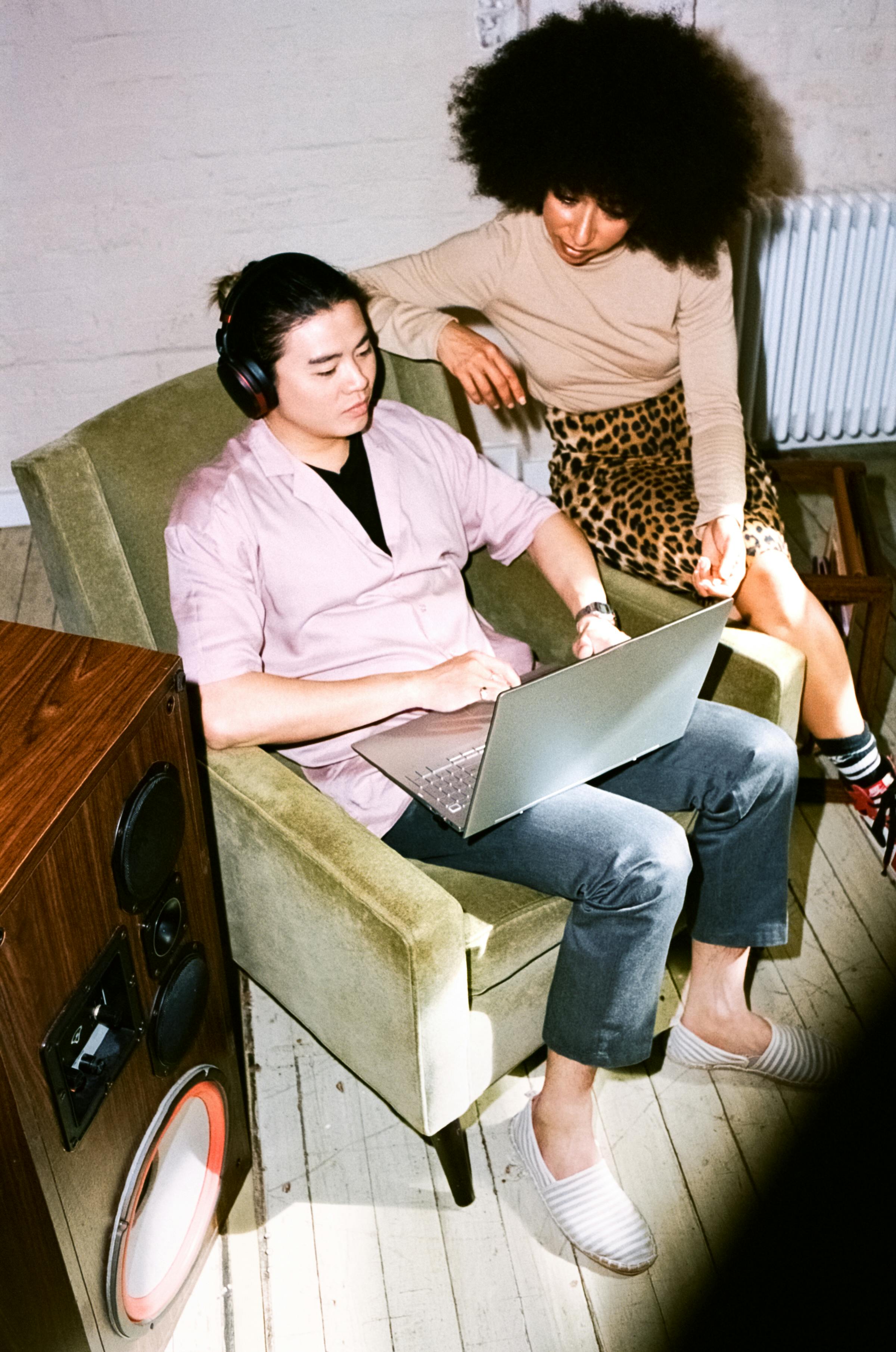 Man in armchair with laptop and headphones, woman leaning over his shoulder to look at the laptop screen with him.