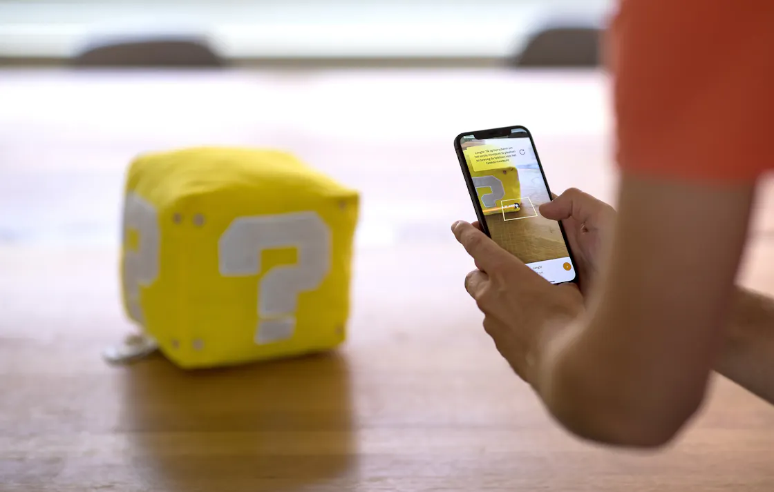 Someone taking a picture of a plush cube with a question mark on it with a phone.